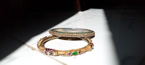 Two ornate bangles are placed on a surface partially illuminated by sunlight. One bangle is slim with green stones embedded around it, while the other is wider with intricate gold designs and multi-colored stones.