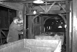 A man operating machinery in an industrial setting appears focused on his work. The surrounding area includes metal structures and equipment, with a cart filled with rock or coal material. The environment is dimly lit, typical of an underground or enclosed industrial facility.