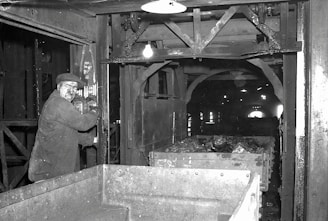 A man operating machinery in an industrial setting appears focused on his work. The surrounding area includes metal structures and equipment, with a cart filled with rock or coal material. The environment is dimly lit, typical of an underground or enclosed industrial facility.