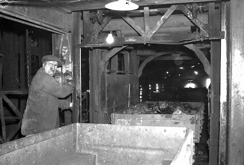 A man operating machinery in an industrial setting appears focused on his work. The surrounding area includes metal structures and equipment, with a cart filled with rock or coal material. The environment is dimly lit, typical of an underground or enclosed industrial facility.