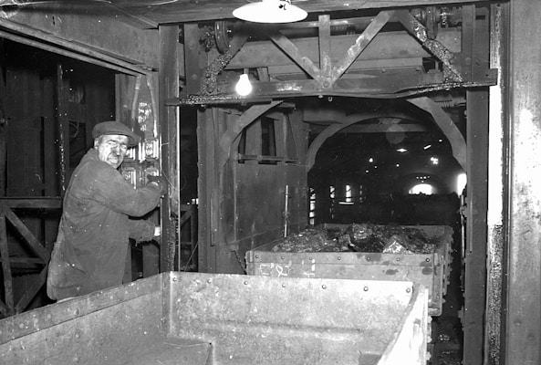 A man operating machinery in an industrial setting appears focused on his work. The surrounding area includes metal structures and equipment, with a cart filled with rock or coal material. The environment is dimly lit, typical of an underground or enclosed industrial facility.