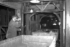 A man operating machinery in an industrial setting appears focused on his work. The surrounding area includes metal structures and equipment, with a cart filled with rock or coal material. The environment is dimly lit, typical of an underground or enclosed industrial facility.