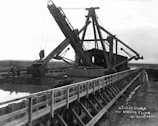 A large dredging machine is positioned on a wooden flume over a waterway, with expansive open land in the background. The structure features long arm mechanisms and multiple pulleys. There are two people visible near the machine, indicating its industrial use.
