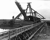 A large dredging machine is positioned on a wooden flume over a waterway, with expansive open land in the background. The structure features long arm mechanisms and multiple pulleys. There are two people visible near the machine, indicating its industrial use.