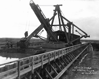 A large dredging machine is positioned on a wooden flume over a waterway, with expansive open land in the background. The structure features long arm mechanisms and multiple pulleys. There are two people visible near the machine, indicating its industrial use.
