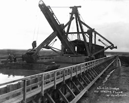 A large dredging machine is positioned on a wooden flume over a waterway, with expansive open land in the background. The structure features long arm mechanisms and multiple pulleys. There are two people visible near the machine, indicating its industrial use.