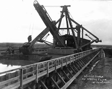 A large dredging machine is positioned on a wooden flume over a waterway, with expansive open land in the background. The structure features long arm mechanisms and multiple pulleys. There are two people visible near the machine, indicating its industrial use.