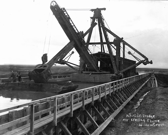 A large dredging machine is positioned on a wooden flume over a waterway, with expansive open land in the background. The structure features long arm mechanisms and multiple pulleys. There are two people visible near the machine, indicating its industrial use.