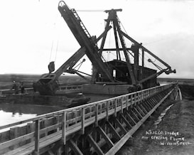A large dredging machine is positioned on a wooden flume over a waterway, with expansive open land in the background. The structure features long arm mechanisms and multiple pulleys. There are two people visible near the machine, indicating its industrial use.
