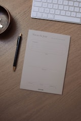 A wooden desk holds a pen with a gold tip, a notepad titled 'Focus du Jour' for organizing daily tasks, a wireless keyboard, and a small round wooden bowl containing some pins.