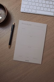A wooden desk holds a pen with a gold tip, a notepad titled 'Focus du Jour' for organizing daily tasks, a wireless keyboard, and a small round wooden bowl containing some pins.