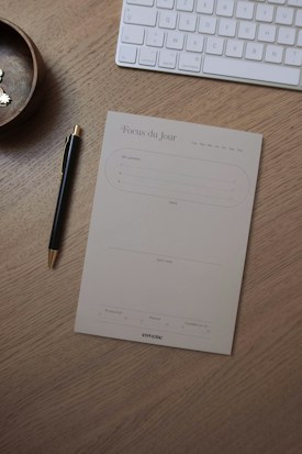A wooden desk holds a pen with a gold tip, a notepad titled 'Focus du Jour' for organizing daily tasks, a wireless keyboard, and a small round wooden bowl containing some pins.