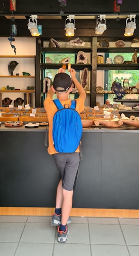 A young boy with a blue backpack and an orange shirt is standing in front of a display of minerals and geodes. The shelves behind the counter are filled with various rocks and crystals, neatly arranged. The environment is well-lit with spotlights overhead, and there is a sense of curiosity as the child reaches for one of the displayed items.