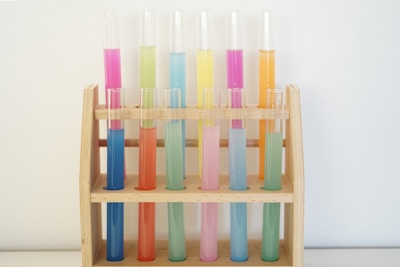 A wooden rack holds a series of test tubes filled with various brightly colored liquids, arranged in a visually striking pattern. The hues range from pink, yellow, blue, and green, creating a vibrant array against a neutral background.