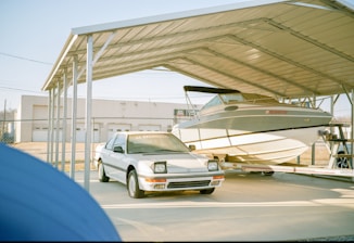 A white car and a boat are parked under a large metal carport. The car, with visible headlights and a Honda logo on its windshield, is in front of the boat. The boat is mounted on a trailer. In the background, a fenced area and a building with garage doors can be seen.