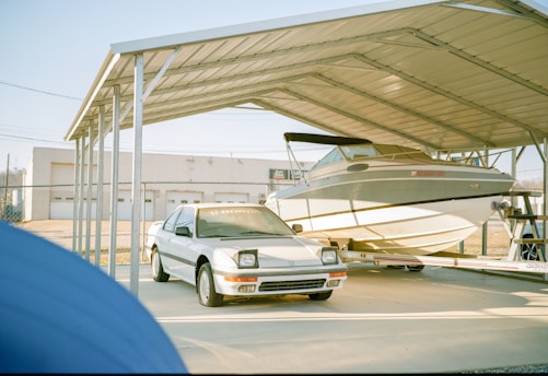 A white car and a boat are parked under a large metal carport. The car, with visible headlights and a Honda logo on its windshield, is in front of the boat. The boat is mounted on a trailer. In the background, a fenced area and a building with garage doors can be seen.