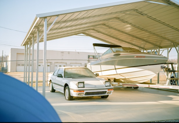 A white car and a boat are parked under a large metal carport. The car, with visible headlights and a Honda logo on its windshield, is in front of the boat. The boat is mounted on a trailer. In the background, a fenced area and a building with garage doors can be seen.