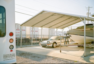 A covered parking area shelters a car and a boat on a trailer. Nearby, a white bus with visible text is partially in view. In the background, a chain-link fence separates the area from a row of parked trailers. The setting appears to be a storage facility or lot, with clear skies and sunlight illuminating the scene.