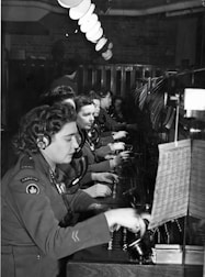 A group of women dressed in military uniforms are operating a vintage telephone switchboard. They are wearing headsets and are focused on connecting calls. The setting appears to be a busy communication center, with rows of desks and lighting fixtures overhead.