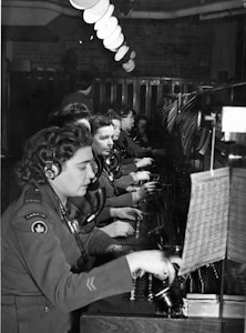 A group of women dressed in military uniforms are operating a vintage telephone switchboard. They are wearing headsets and are focused on connecting calls. The setting appears to be a busy communication center, with rows of desks and lighting fixtures overhead.
