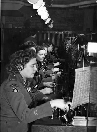A group of women dressed in military uniforms are operating a vintage telephone switchboard. They are wearing headsets and are focused on connecting calls. The setting appears to be a busy communication center, with rows of desks and lighting fixtures overhead.
