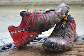 A pair of muddy red and black trail running shoes with prominent logo and design elements are placed on a doormat. The shoes are heavily soiled, suggesting they were used in a rugged outdoor environment. The background shows a neutral, blurred surface.