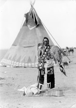 A person stands in traditional attire in front of a large teepee. The clothing features intricate patterns and decorations, including feathers. The background includes additional tents and a dry, open landscape.