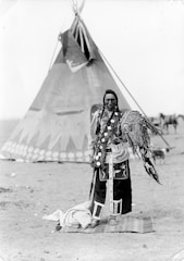 A person stands in traditional attire in front of a large teepee. The clothing features intricate patterns and decorations, including feathers. The background includes additional tents and a dry, open landscape.