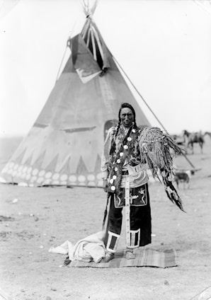 A person stands in traditional attire in front of a large teepee. The clothing features intricate patterns and decorations, including feathers. The background includes additional tents and a dry, open landscape.