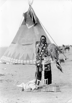 A person stands in traditional attire in front of a large teepee. The clothing features intricate patterns and decorations, including feathers. The background includes additional tents and a dry, open landscape.