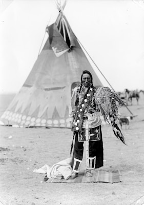A person stands in traditional attire in front of a large teepee. The clothing features intricate patterns and decorations, including feathers. The background includes additional tents and a dry, open landscape.