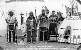 Four Indigenous people are standing in traditional clothing in front of a decorated teepee. Two men are wearing feathered headdresses and holding staffs. A woman and an elder are also present, with the woman wearing a dress adorned with beadwork. The background shows other tents or teepees and what appears to be a wagon wheel, suggesting a cultural gathering or event.