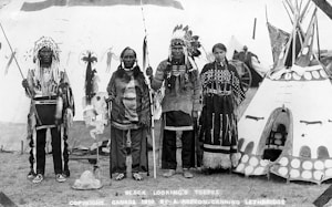 Four Indigenous people are standing in traditional clothing in front of a decorated teepee. Two men are wearing feathered headdresses and holding staffs. A woman and an elder are also present, with the woman wearing a dress adorned with beadwork. The background shows other tents or teepees and what appears to be a wagon wheel, suggesting a cultural gathering or event.