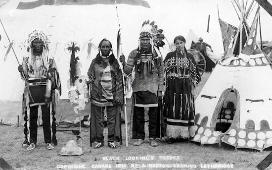 Four Indigenous people are standing in traditional clothing in front of a decorated teepee. Two men are wearing feathered headdresses and holding staffs. A woman and an elder are also present, with the woman wearing a dress adorned with beadwork. The background shows other tents or teepees and what appears to be a wagon wheel, suggesting a cultural gathering or event.
