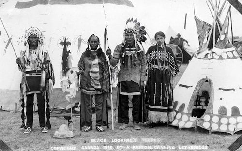 Four Indigenous people are standing in traditional clothing in front of a decorated teepee. Two men are wearing feathered headdresses and holding staffs. A woman and an elder are also present, with the woman wearing a dress adorned with beadwork. The background shows other tents or teepees and what appears to be a wagon wheel, suggesting a cultural gathering or event.