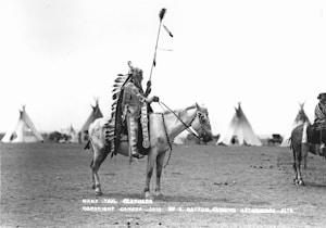 A person in traditional indigenous attire on horseback, with a headdress adorned with feathers and a decorated robe. The background shows a series of teepees on an open plain under a clear sky.