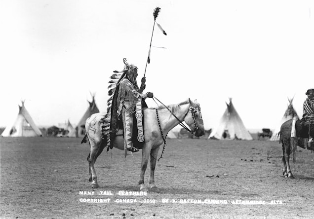 A person in traditional indigenous attire on horseback, with a headdress adorned with feathers and a decorated robe. The background shows a series of teepees on an open plain under a clear sky.