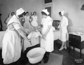 A group of nurses wearing traditional uniforms are gathered around a hospital bed in a clinical setting. One nurse appears to be changing linens or adjusting the bedding of a patient. A basin is placed on the floor, and there is a small table with medical tools and supplies nearby. The setting suggests a hospital or medical training environment from a previous era.