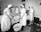 A group of nurses wearing traditional uniforms are gathered around a hospital bed in a clinical setting. One nurse appears to be changing linens or adjusting the bedding of a patient. A basin is placed on the floor, and there is a small table with medical tools and supplies nearby. The setting suggests a hospital or medical training environment from a previous era.
