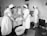 A group of nurses wearing traditional uniforms are gathered around a hospital bed in a clinical setting. One nurse appears to be changing linens or adjusting the bedding of a patient. A basin is placed on the floor, and there is a small table with medical tools and supplies nearby. The setting suggests a hospital or medical training environment from a previous era.