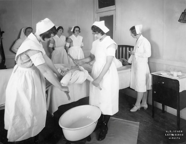 A group of nurses wearing traditional uniforms are gathered around a hospital bed in a clinical setting. One nurse appears to be changing linens or adjusting the bedding of a patient. A basin is placed on the floor, and there is a small table with medical tools and supplies nearby. The setting suggests a hospital or medical training environment from a previous era.