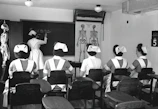 A classroom with five nurses in uniforms seated at desks, facing a blackboard where another nurse is writing. Anatomical charts and a skeleton model are visible, indicating a lesson on human anatomy.