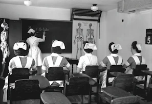 A classroom with five nurses in uniforms seated at desks, facing a blackboard where another nurse is writing. Anatomical charts and a skeleton model are visible, indicating a lesson on human anatomy.