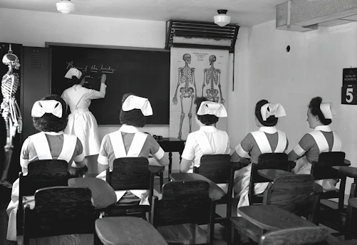 A classroom with five nurses in uniforms seated at desks, facing a blackboard where another nurse is writing. Anatomical charts and a skeleton model are visible, indicating a lesson on human anatomy.