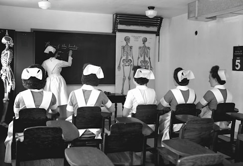 A classroom with five nurses in uniforms seated at desks, facing a blackboard where another nurse is writing. Anatomical charts and a skeleton model are visible, indicating a lesson on human anatomy.