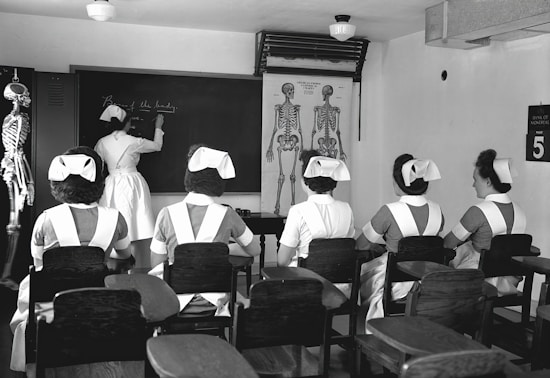 A classroom with five nurses in uniforms seated at desks, facing a blackboard where another nurse is writing. Anatomical charts and a skeleton model are visible, indicating a lesson on human anatomy.