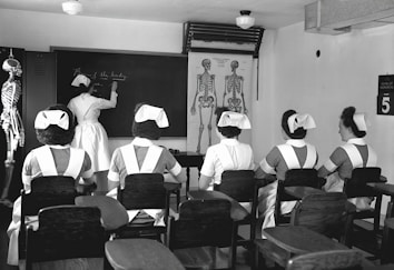 A classroom with five nurses in uniforms seated at desks, facing a blackboard where another nurse is writing. Anatomical charts and a skeleton model are visible, indicating a lesson on human anatomy.
