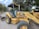 A yellow backhoe loader is parked on a paved road surrounded by trees and greenery. The machine, labeled with '414E IL' and 'Cashman', has large tires and visible interior controls. The sky is partly cloudy.
