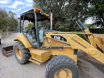 A yellow backhoe loader is parked on a paved road surrounded by trees and greenery. The machine, labeled with '414E IL' and 'Cashman', has large tires and visible interior controls. The sky is partly cloudy.