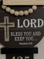A decorative display featuring a wooden cross and a string of large wooden beads. The background has the text 'LORD BLESS YOU AND KEEP YOU... Numbers 6:24' in white, set against a dark circular board.